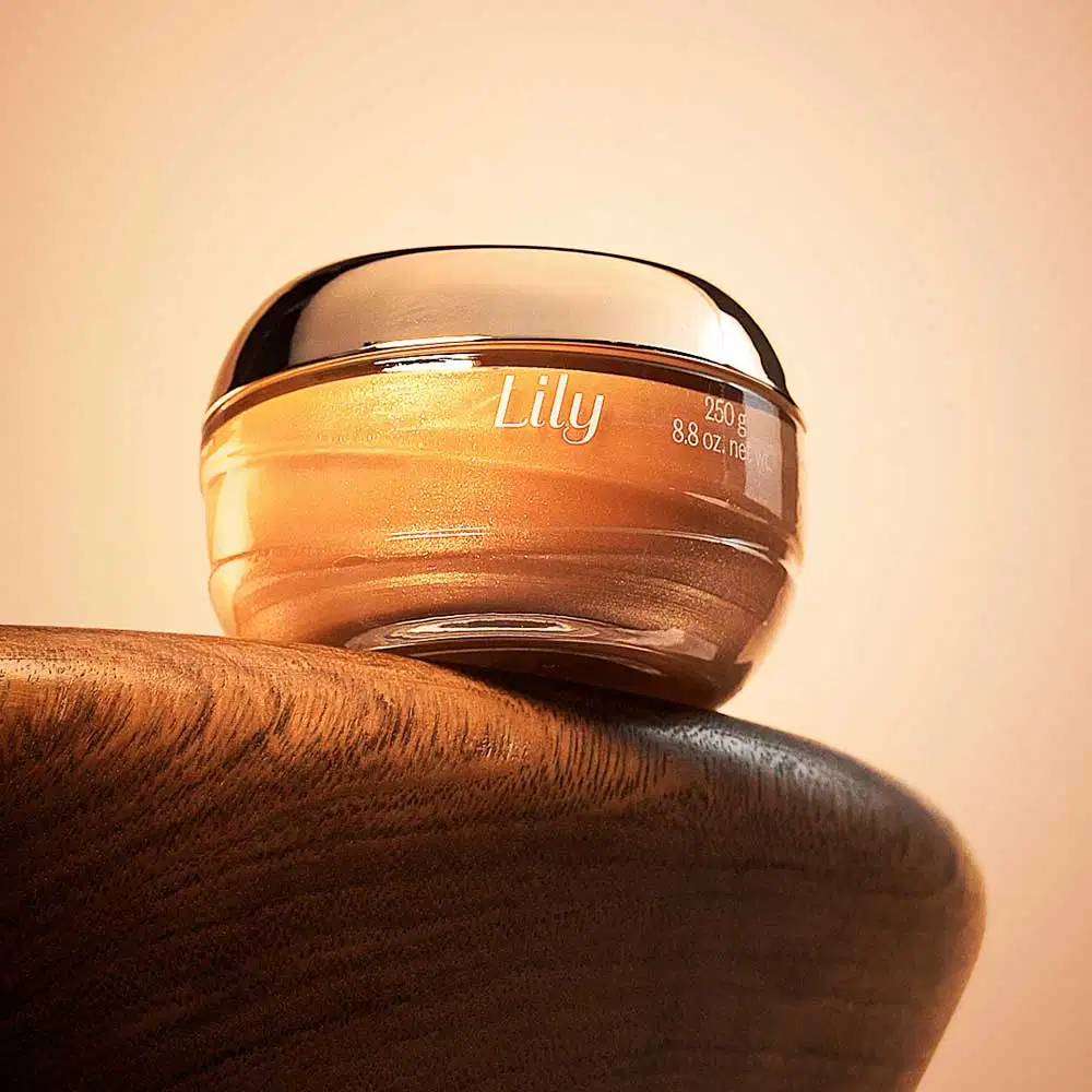 Pote de vidro om creme dourado, tampa dourada e letras bege de geleia iluminadora corporal Lily Glow. posicionado sobre superfície de madeira, com fundo bege claro e iluminação natural que destaca o gel dourado dentro da embalagem com tampa espelhada.
