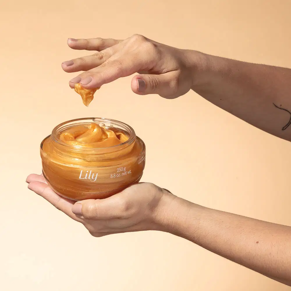Uma mão feminina segura um pote de vidro com creme dourado  e a outra mão passa um dedo no produto, tudo sobre fundo em tons de bege.