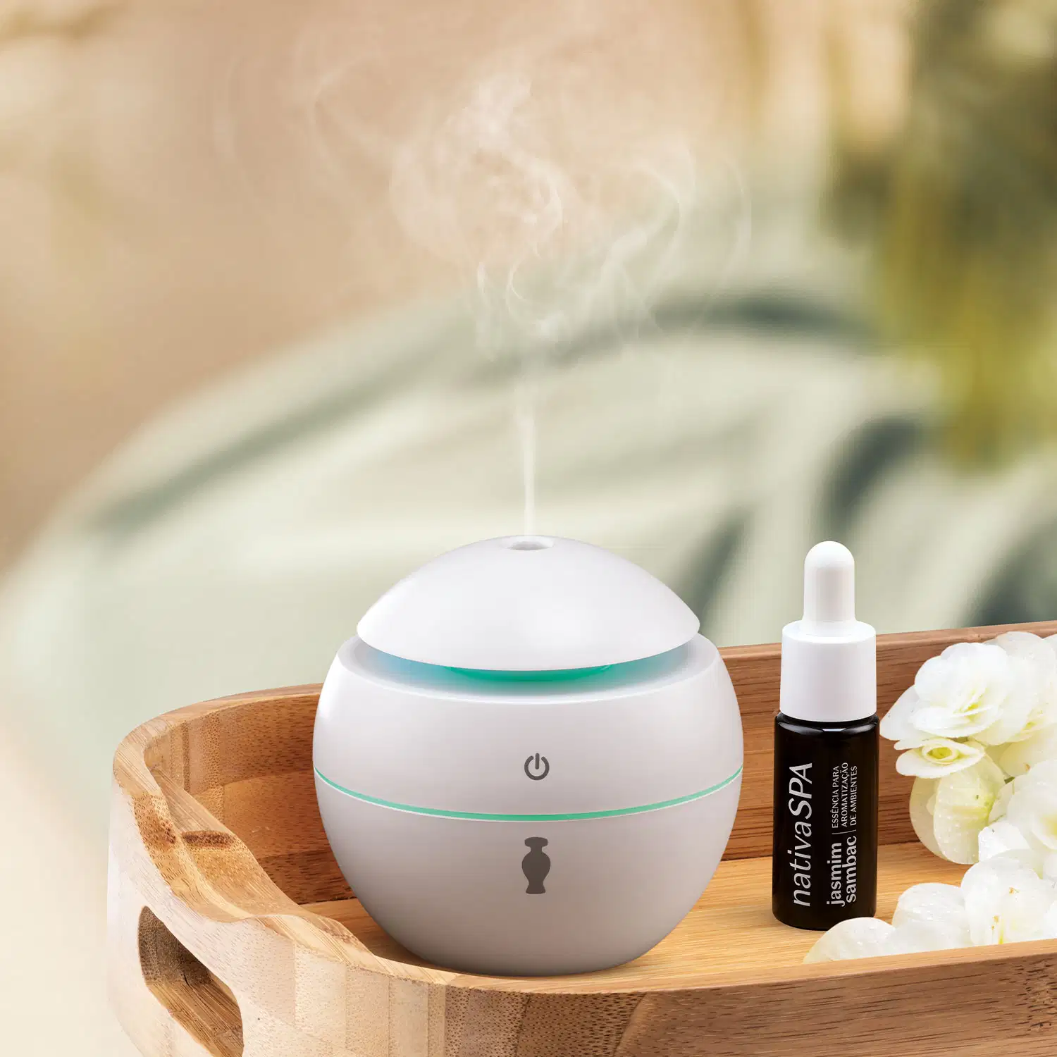 Aromatizador elétrico de ambientes branco com tampa em formato de cúpula, botão power cinza no topo e ícone da marca O Boticário no corpo do pote em bandeja de madeira ao lado de frasco de óleo essencial e fonte ao fundo desfocada.