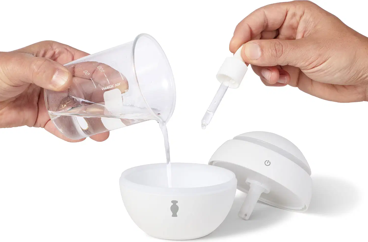 Mão feminina despejando água dentro do pote principal do aromatizador elétrico de ambientes branco. Uma mão masculina pinga essência dentro do mesmo recipiente, enquanto a tampa do aparelho está posicionada ao lado.