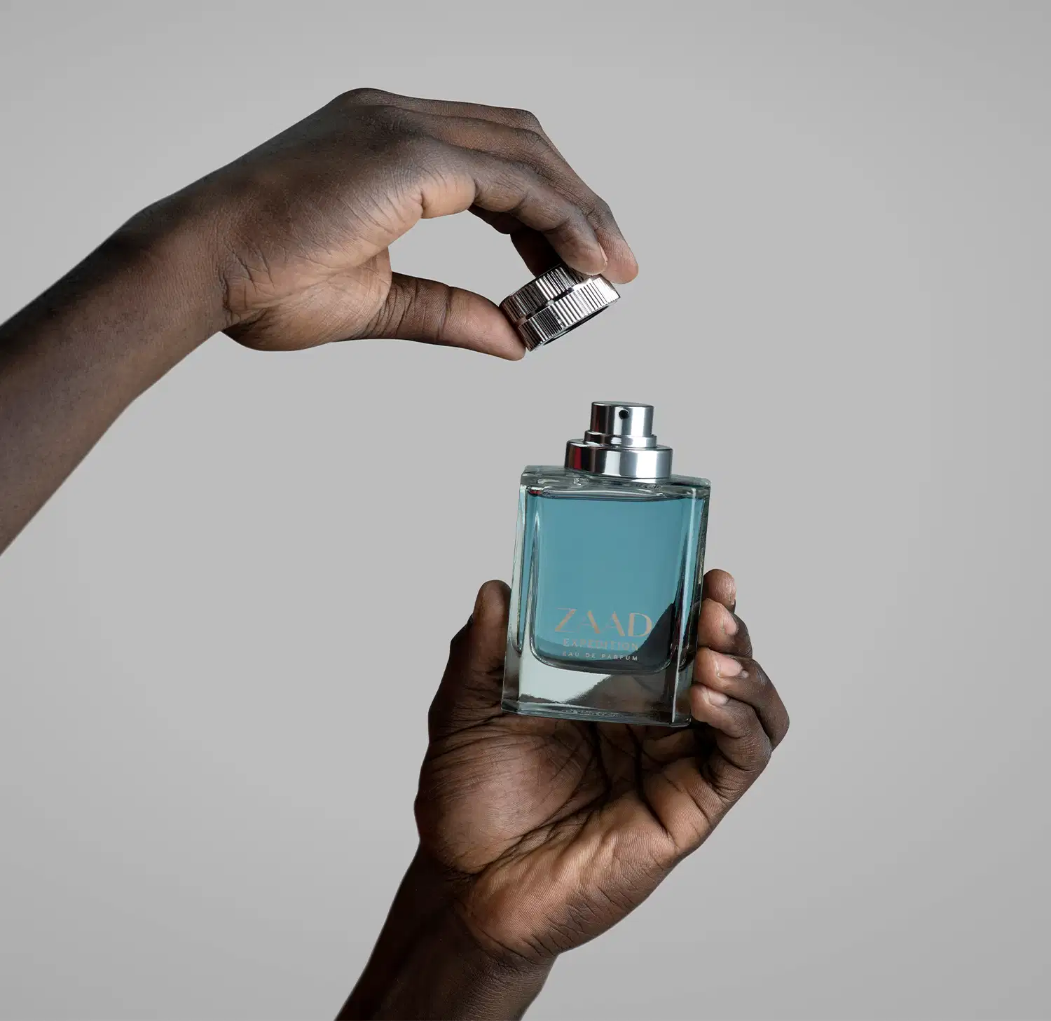 Uma mão masculina segura um Vidro transparente em formato quadrado, com líquido azul acinzentado de Eau de Parfum Zaad Expedition e a outra mão segura a tampa pratada, sobre fundo cinza claro.