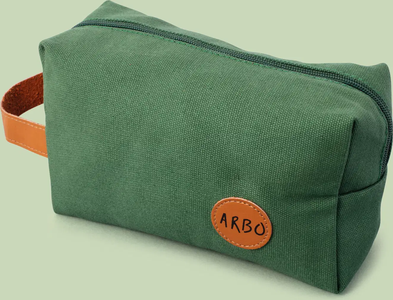 Nécessaire retangular de lona verde com alça de couro caramelo, zíper verde e logo Arbo em patch circular também em couro caramelo sobre fundo cinza.