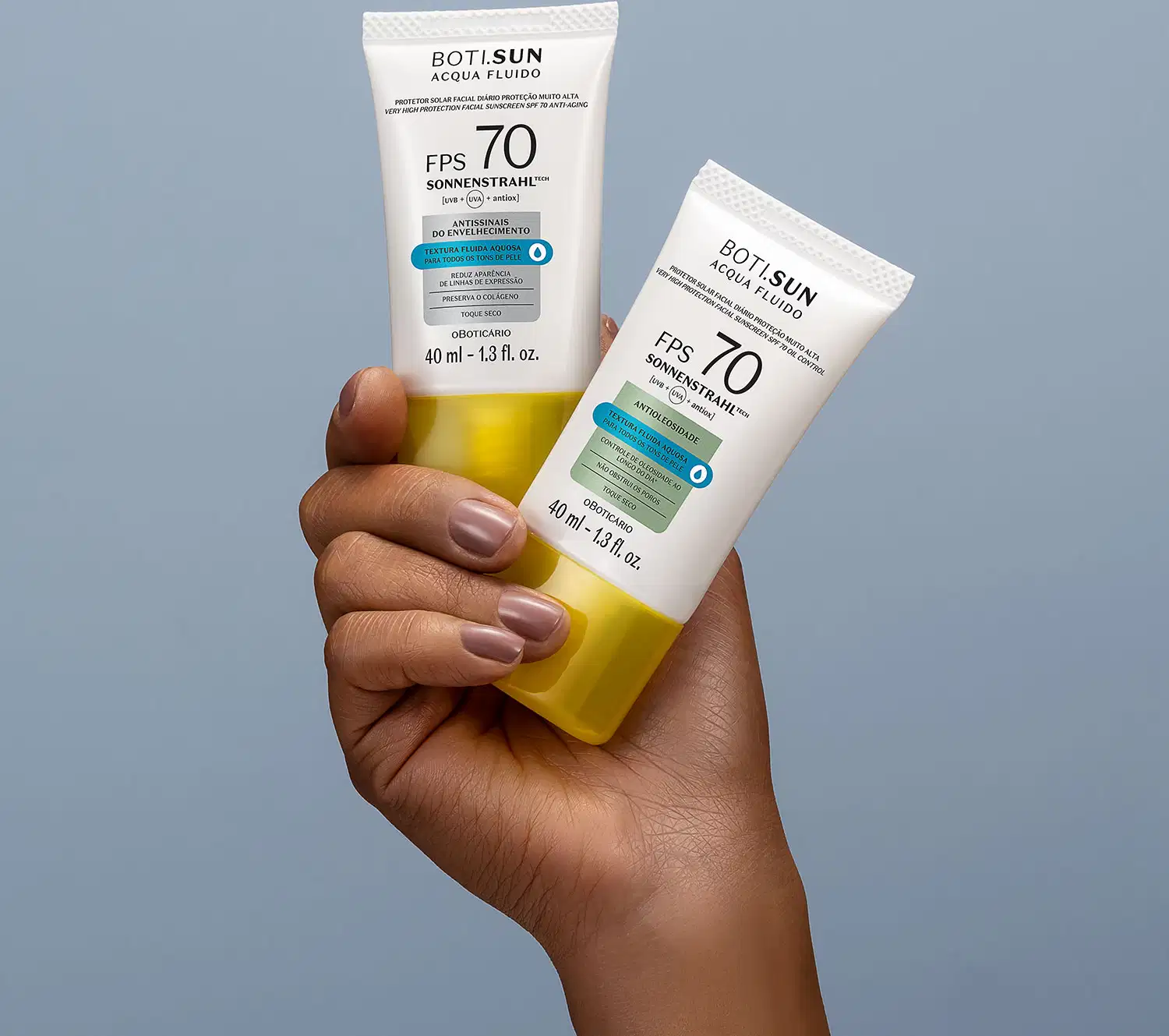 Uma mão feminina segura dois frascos brancos com tampas amarelas, letras pretas e detalhes prata e azul, de protetor solar facial antioleosidade FPS 70 Acqua Fluído da marca Boti.Sun, sobre fundo azul.