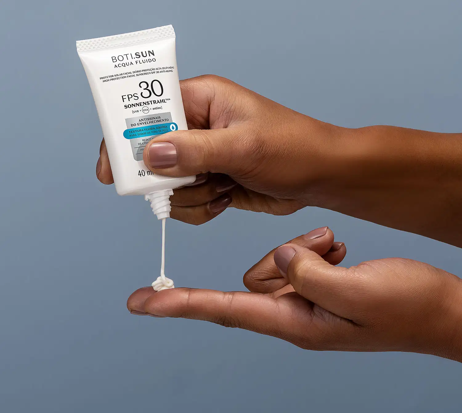 Uma mão feminina segura um frasco branco com letras pretas e detalhes prata e azul de protetor solar facial antissinais FPS 30 Acqua Fluído da marca Boti.Sun e aplica o produto na outra mão, tudo em fundo azul escuro.