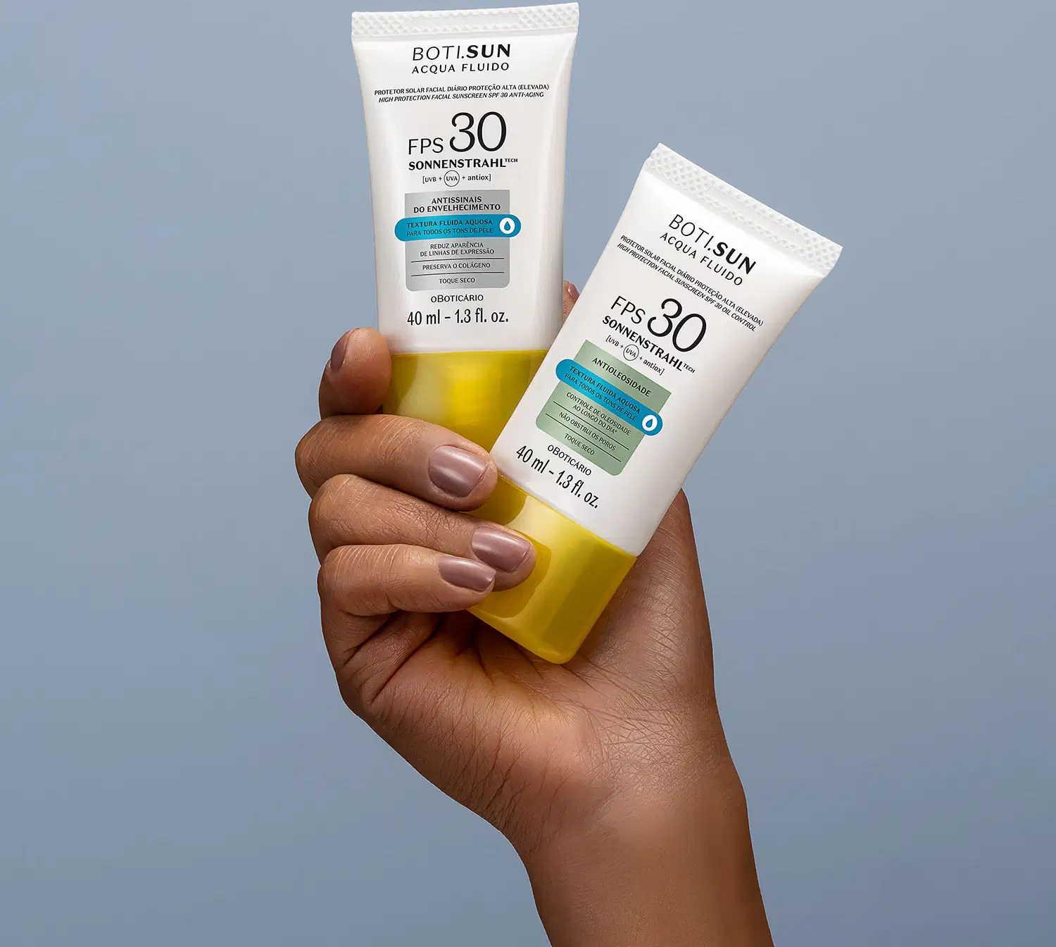 Uma mão feminina segura dois frascos brancos com tampas amarelas, letras pretas e detalhes prata e azul, de protetor solar facial antissinais FPS 30 Acqua Fluído da marca Boti.Sun, sobre fundo azul.