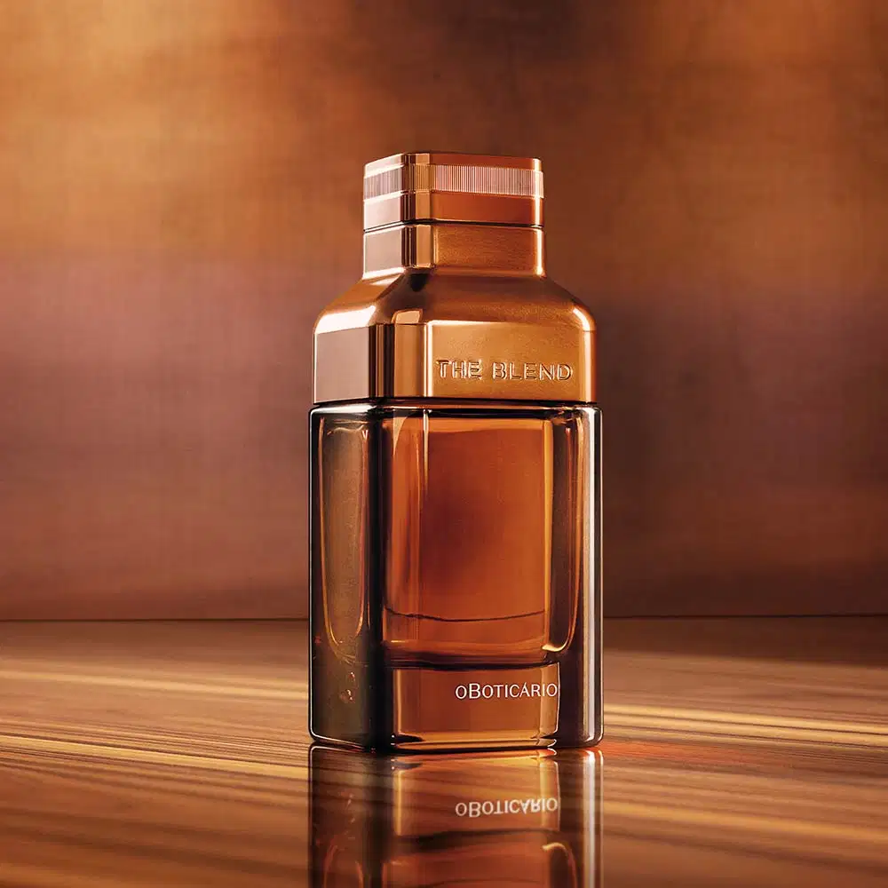 Vidro em tom de marrom fosco com detalhes em marrom metalizado e letras azuis contendo Eau de Parfum The Blend posicionado sobre superfície de madeira clara, com fundo desfocado em tons quentes de marrom e bege, refletindo luz dourada.