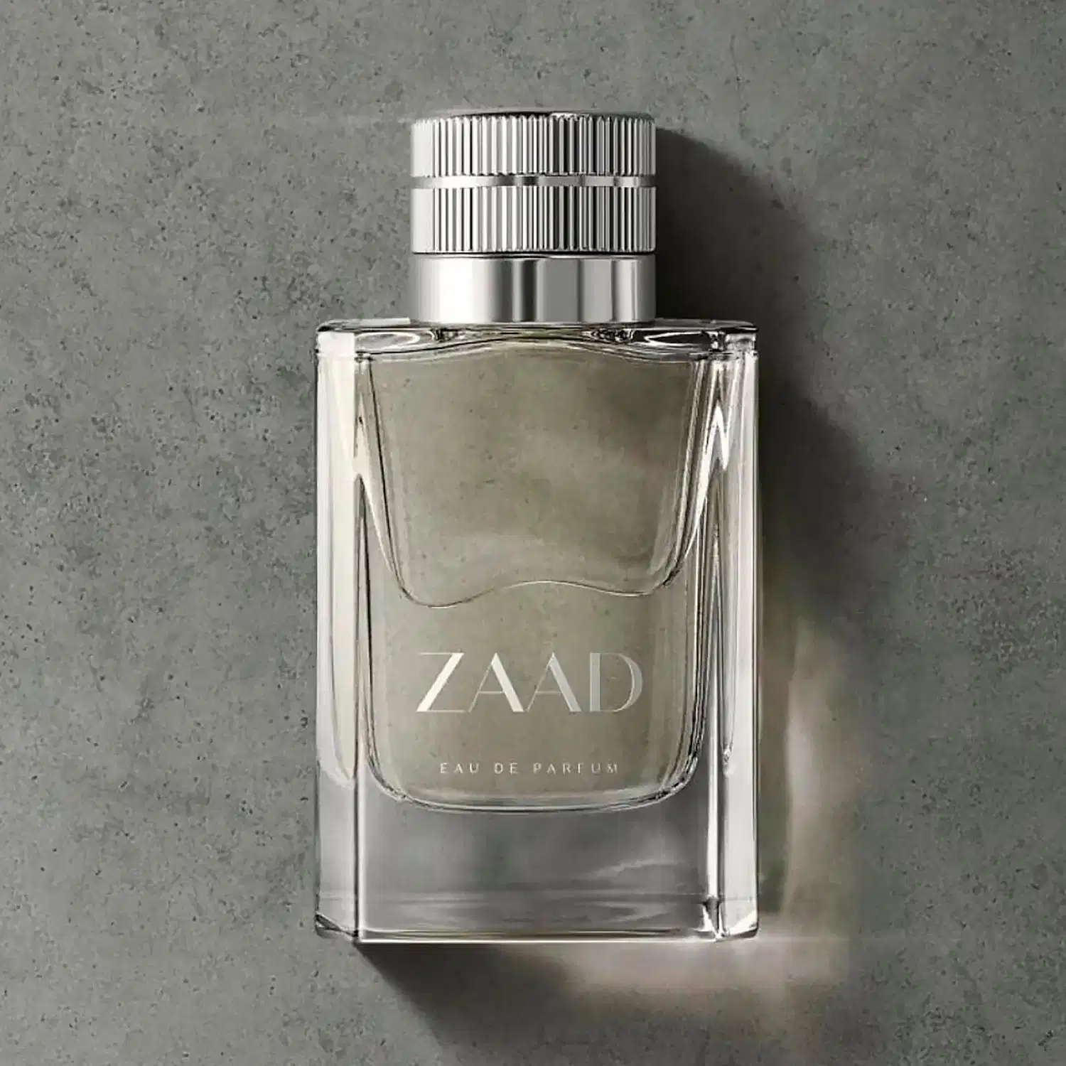 Vidro transparente com líquido cinza, tampa prateada e letras cinzas de Eau de Parfum Zaad, posicionado sobre superfície cinza de concreto com sombra projetada ao lado.