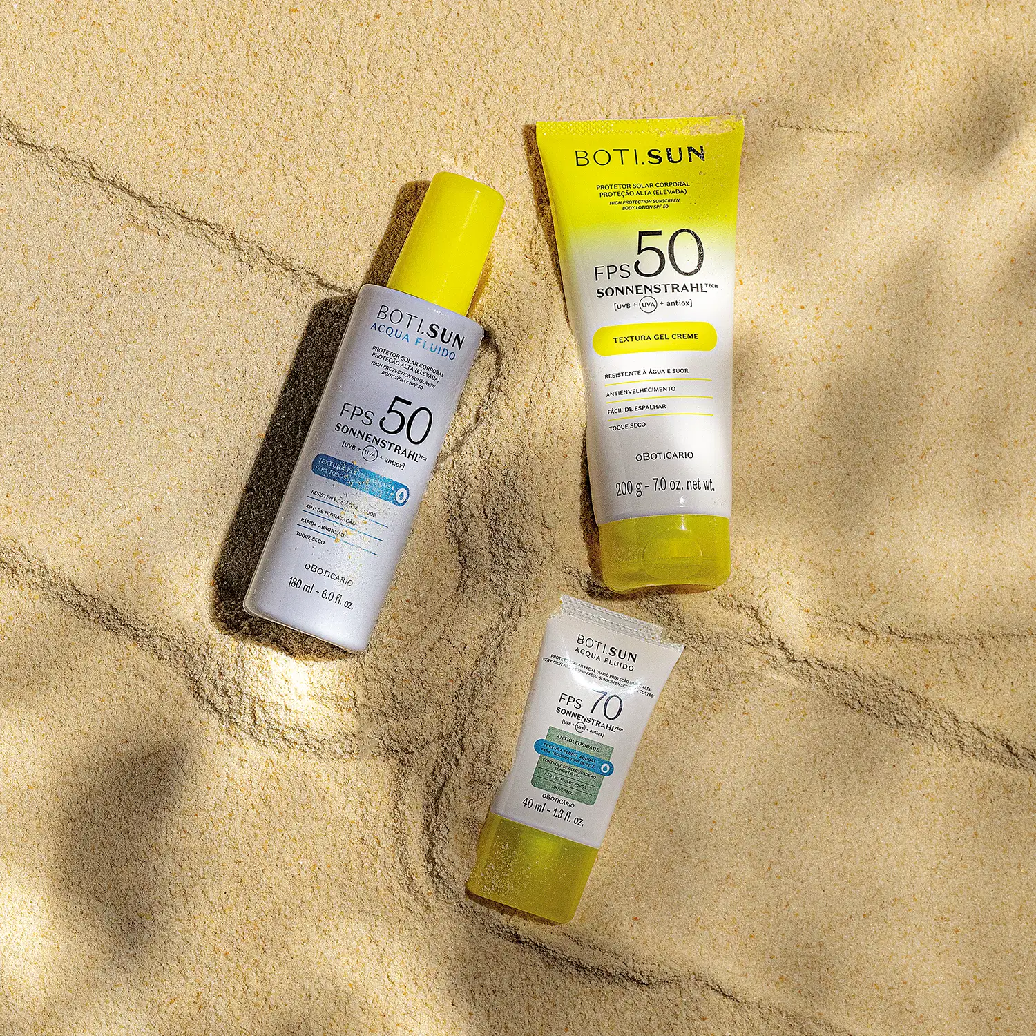 Três produtos Boti.Sun com protetor solar FPS 50 e 70 posicionados sobre areia clara, com sombras de luz natural, incluindo frasco spray, tubo gel-creme e embalagem compacta.
