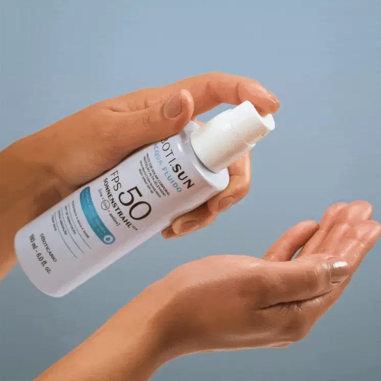Uma mão feminina segura um frasco branco com borrifador também branco e borrifa um pouco do spray protetor solar FPS 50 na outra mão, tudo em um fundo azul.