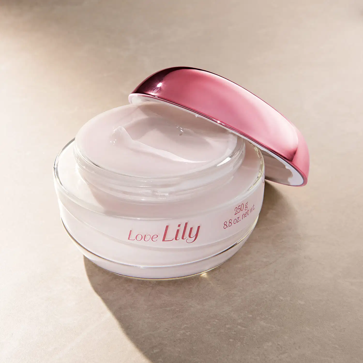 Pote de vidro transparente aberto com creme acetinado hidratante corporal da marca Love Lily na cor branca, tampa roxa encostada ao lado, em fundo lilás com sombra.