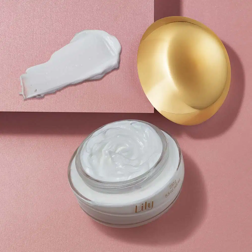 Pote de vidro transparente com conteúdo branco aberto, tampa dourada encostada ao lado e um pequena porção de creme hidratante desodorante corporal da marca Lily espalhada na superfície bege.