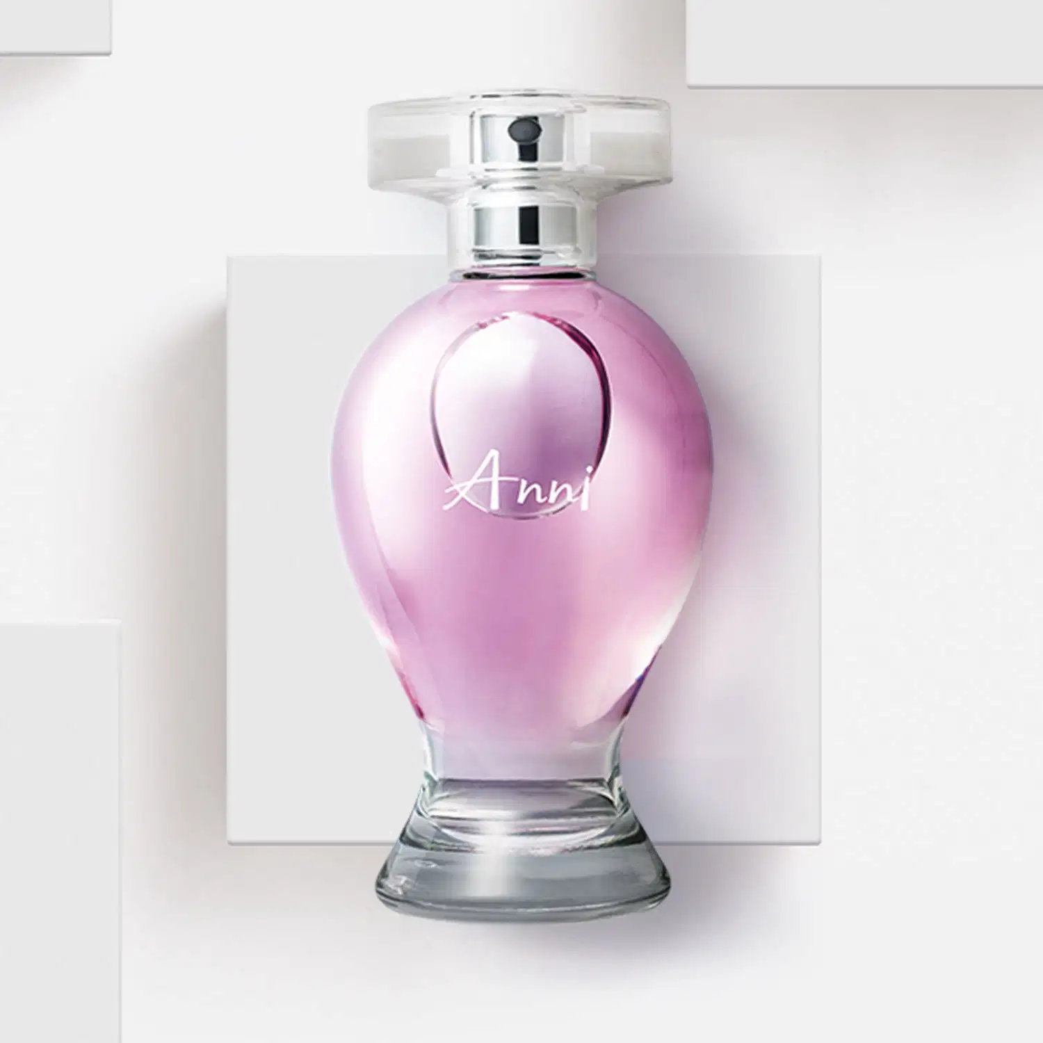 Vidro curvilíneo transparente com liquido rosa, pulverizador prateado e tampa cristal contendo desodorante colônia Boticollection Anni, posicionado sobre superfície branca com painel geométrico ao fundo em ambiente minimalista.