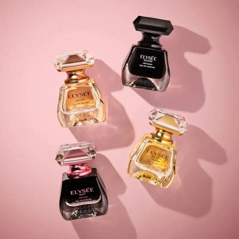 Quatro frascos de itens de perfumaria Elysée em tons dourado, rosa e preto com tampas geométricas, dispostos sobre fundo rosa claro com sombras projetadas.