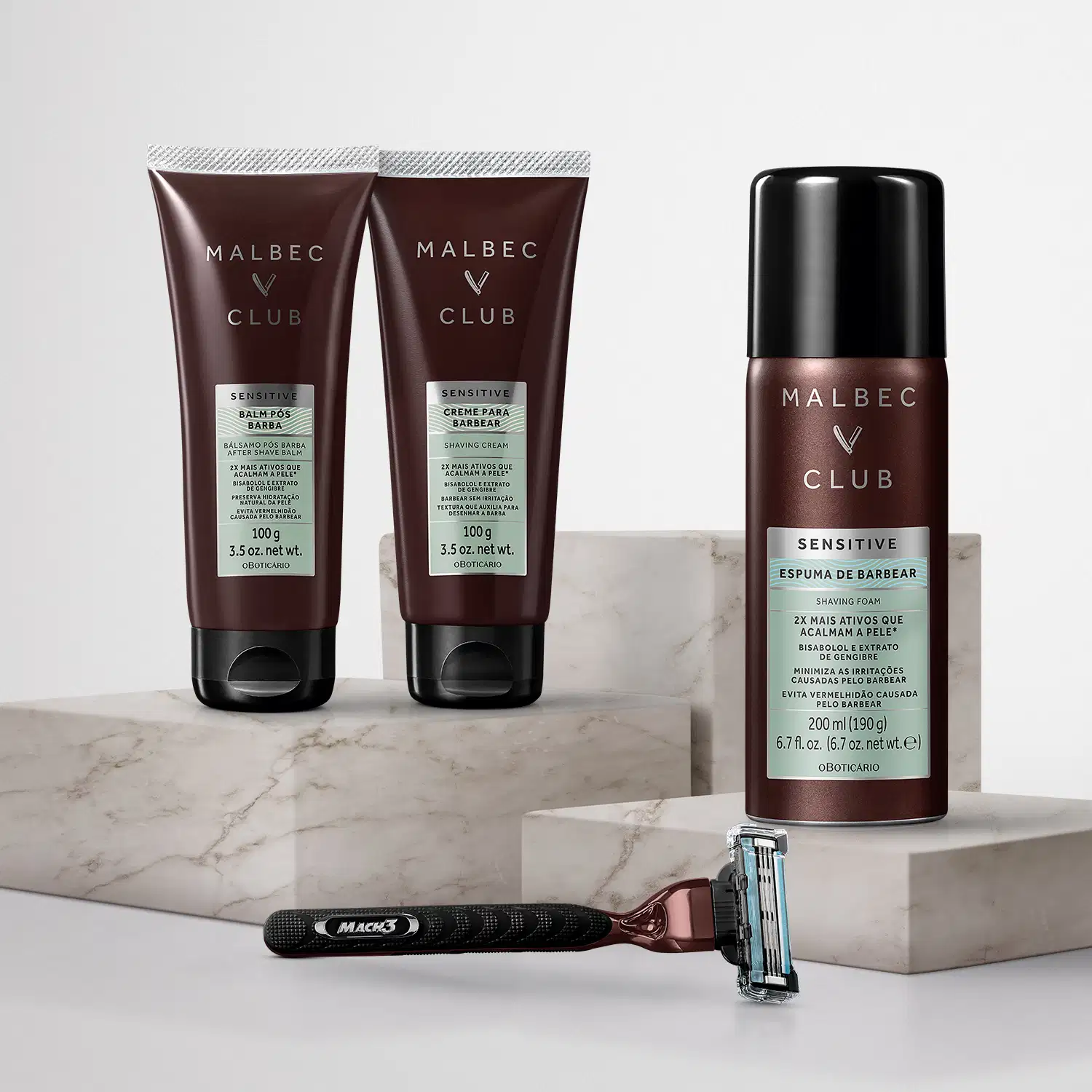 Dois tubos de creme para barbear da marca Malbec Club, em embalagem marrom escura com texto branco. Também há um frasco de espuma de barbear da mesma marca, em embalagem verde clara. Junto, um aparelho de barbear preto com cabo.