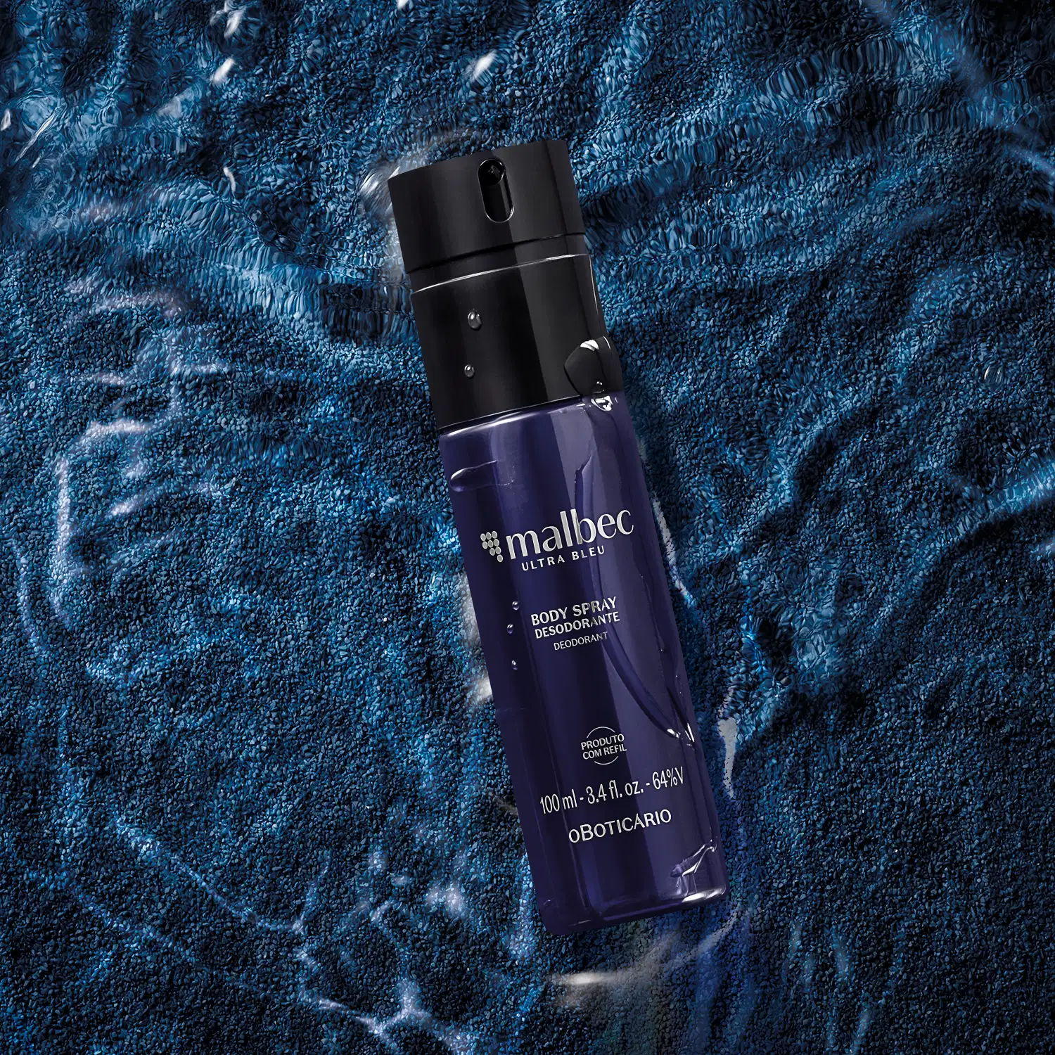 Frasco azul escuro com letras cinzas contendo 100ml de body spray Malbec Ultra Bleu, posicionado sobre superfície azul com textura de fios e padrões geométricos, com partículas brancas dispersas ao redor.