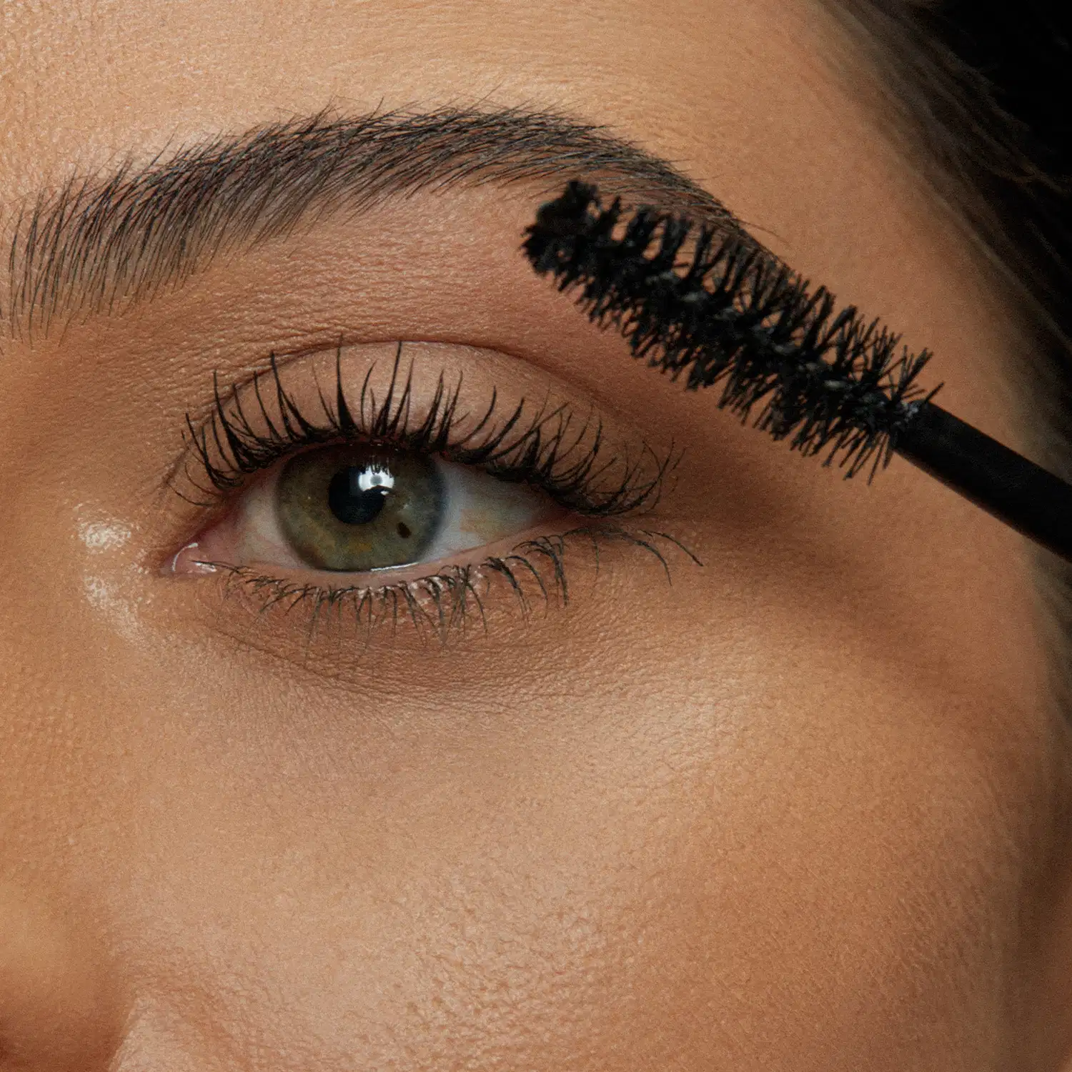 Close-up de um rosto feminino evidenciando os olhos, onde uma escova aplicadora aplica a máscara de cílios Make B. Efeito Cílios Postiços nos cílios, deixando os fios alongados e bem definidos.