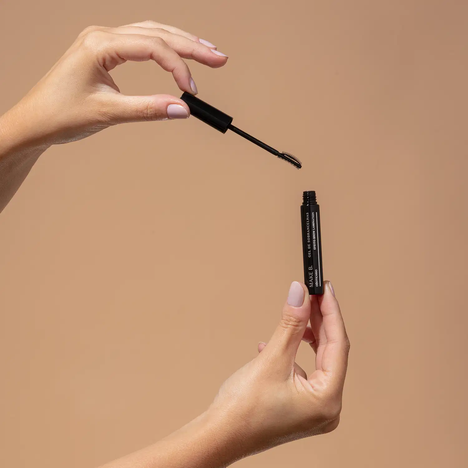 Uma mão feminina segura um tubo preto de máscara de sobrancelhas em gel Make.B Brow Lamination e a outra mão segura a tampa com a haste e a escova aplicadora pretas, sobre fundo em tons de bege.