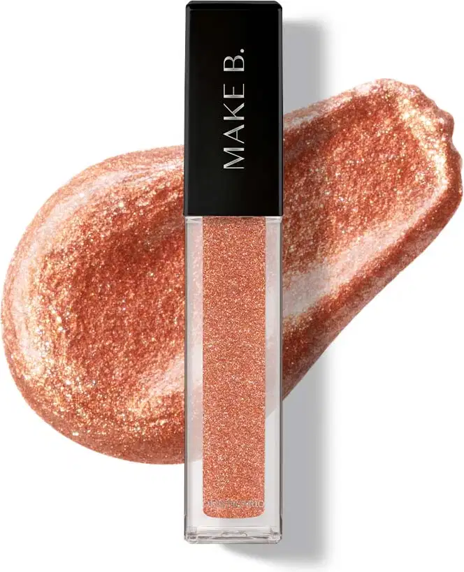 Batom líquido Diamond Dourado Luxury da marca Make B. em tubo retangular transparente, produto de cor dourada, tampa e haste pretas e esponja aplicadora macia., apresentado  textura sobre manchas do produto aplicado em superfície branca.