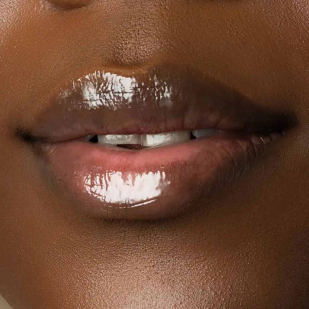 Close-up de um rosto feminino de pele negra evidenciando os lábios brilhantes que usam o gloss labial Incolor Crystal Icon da marca Make B. 