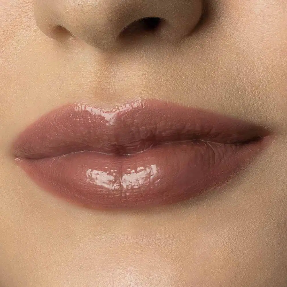 Close-up de um rosto feminino de pele branca evidenciando os lábios brilhantes e rosados que usam o gloss labial Marrom Claro Soft Silk da marca Make B. 