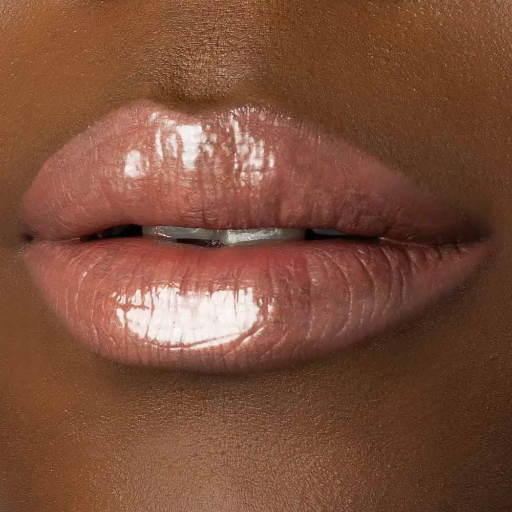 Close-up de um rosto feminino de pele negra evidenciando os lábios brilhantes e rosados que usam o gloss labial Marrom Claro Soft Silk da marca Make B. 