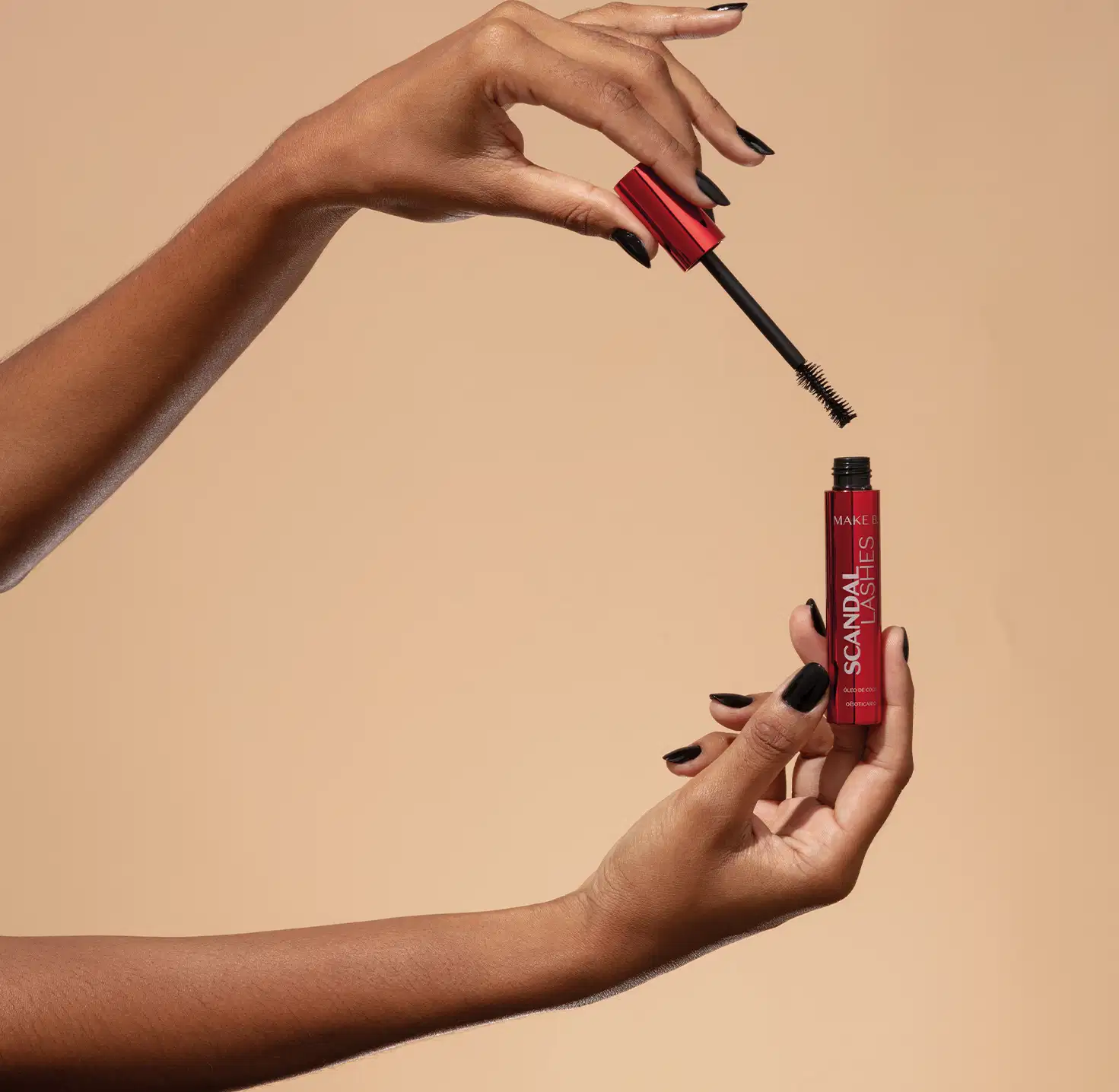 Uma mão feminina segura um tubo vermelho metalizado com letras azuis de máscara de cílios Make B. Scandal Lashes e a outra mão segura a tampa vermelha metalizada com haste e aplicador pretos, sobre fundo em tons de bege.