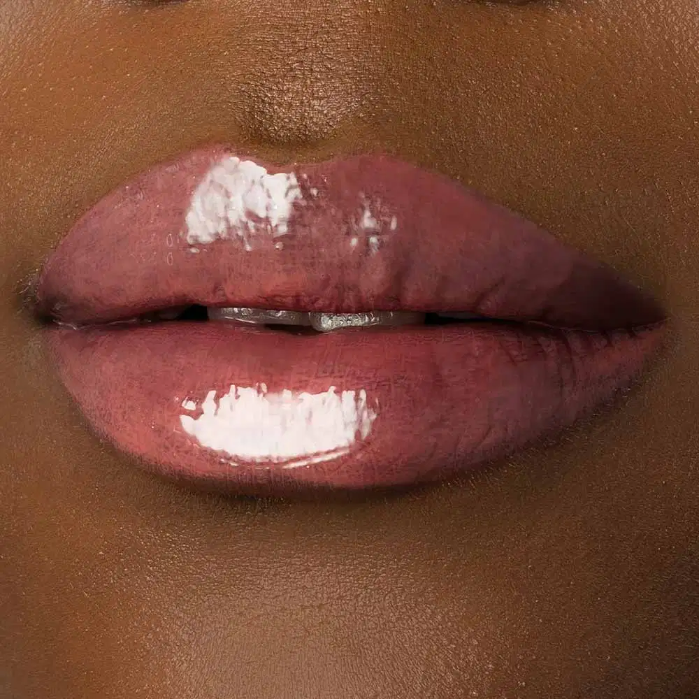 Close-up de um rosto feminino de pele negra evidenciando os lábios brilhantes e rosados que usam o gloss labial Rosa New Rose da marca Make B. 