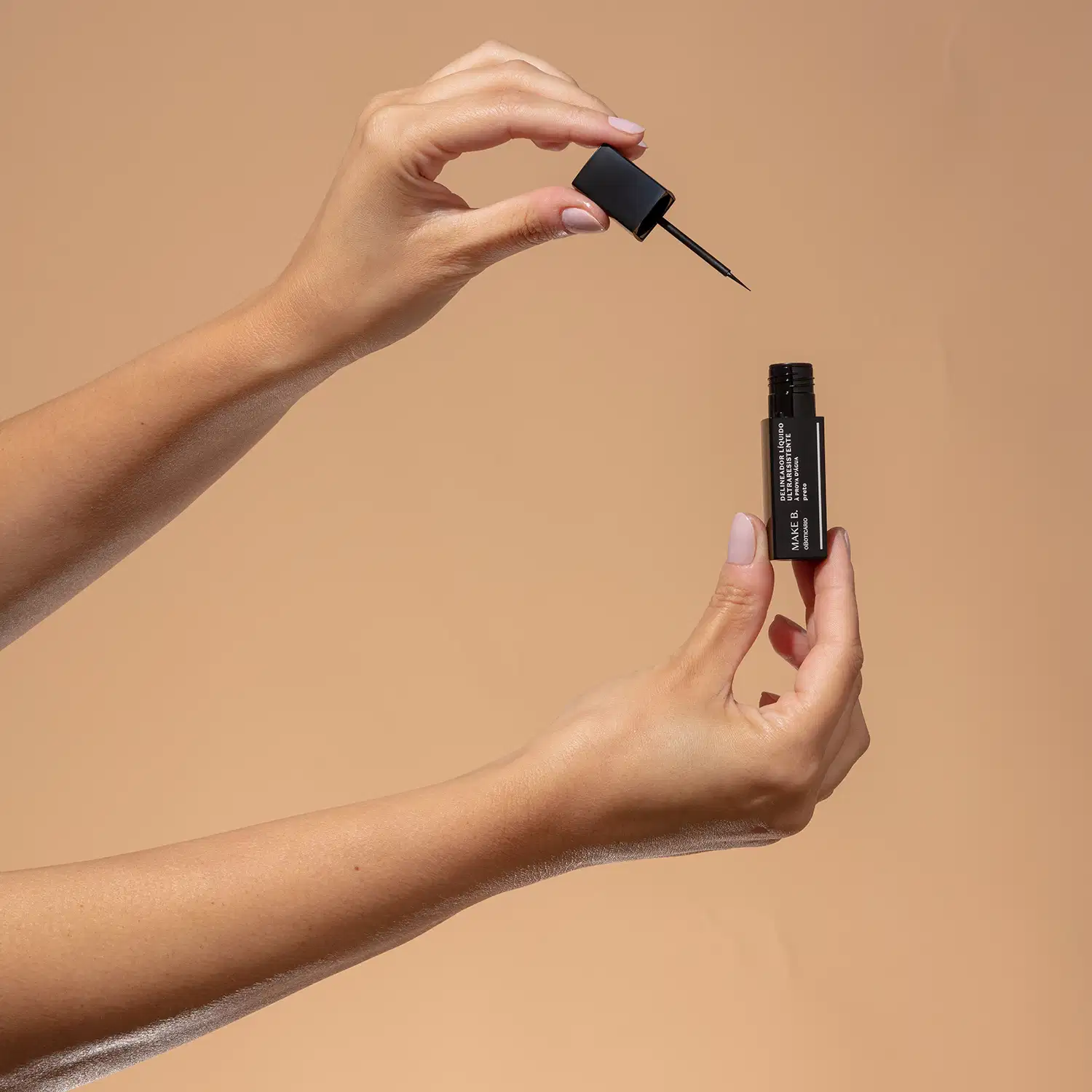 Uma mão feminina segura um tubo preto  com letras brancas de delineador líquido para olhos na cor preta da marca Make B., e a outra mão segura a tampa com haste e aplicador fino, tudo sobre fundo bege.