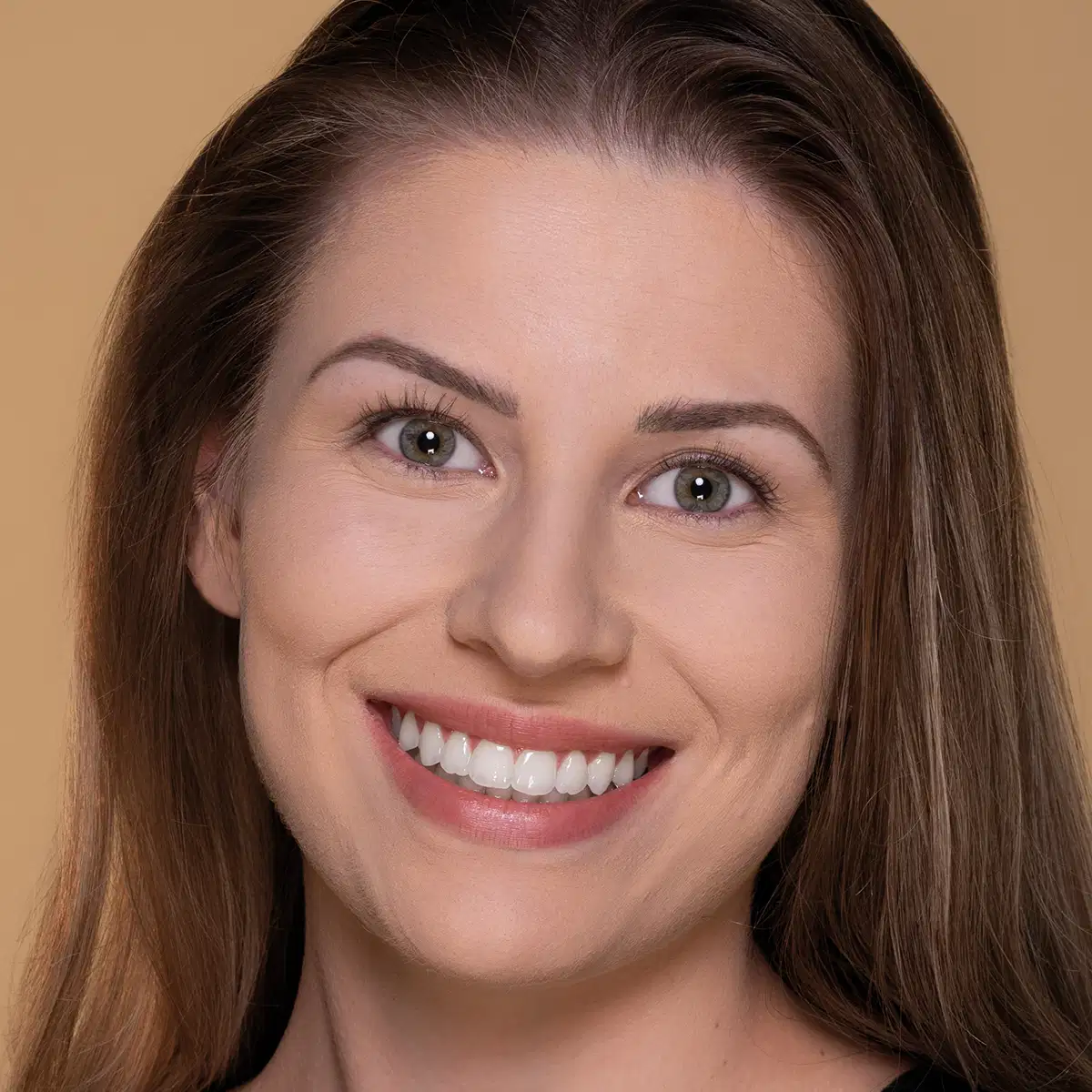 Mulher branca de cabelos castanhos lisos sorrindo com dentes brancos e lábios rosados, posada contra fundo bege liso; não há produto de maquiagem, marca ou embalagem visível na imagem.