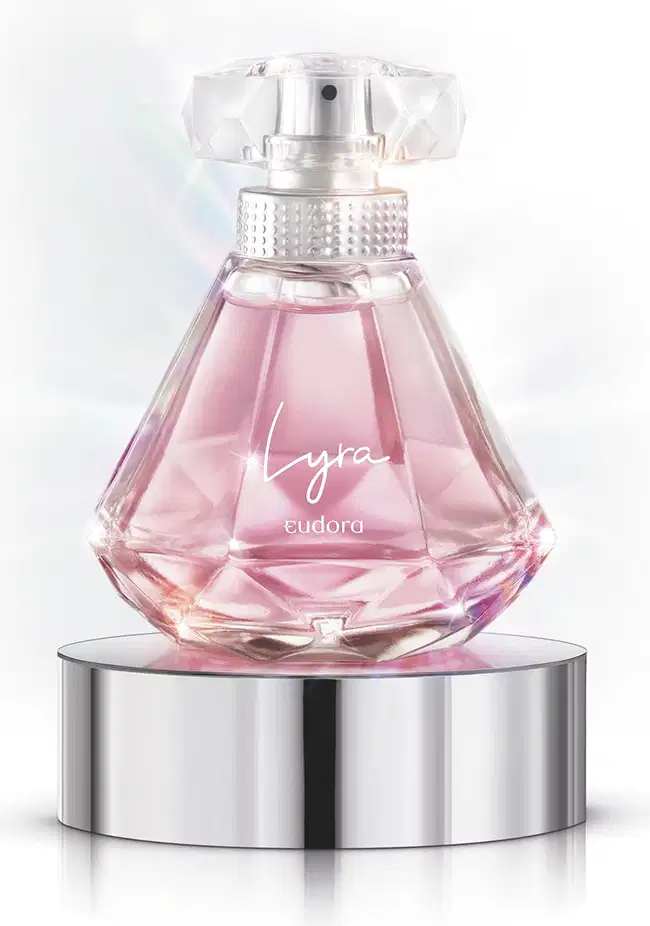 A imagem mostra o Desodorante Colônia Lyra, da Eudora, em frasco de vidro transparente com formato geométrico facetado. O líquido dentro é de cor rosa claro. O frasco repousa sobre uma base cilíndrica de metal prateado.