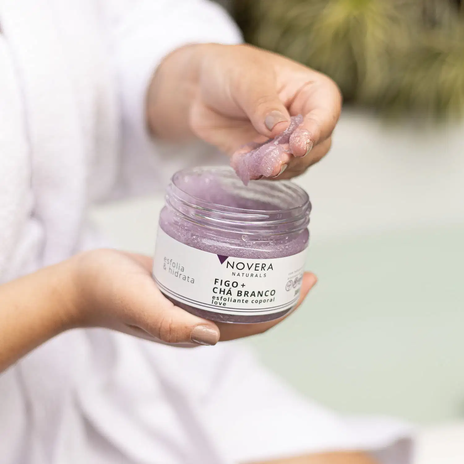 A imagem mostra duas mãos segurando um pequeno pote de vidro transparente contendo um produto corporal esfoliante natural. O produto tem uma cor roxa clara e parece ter uma textura cremosa.