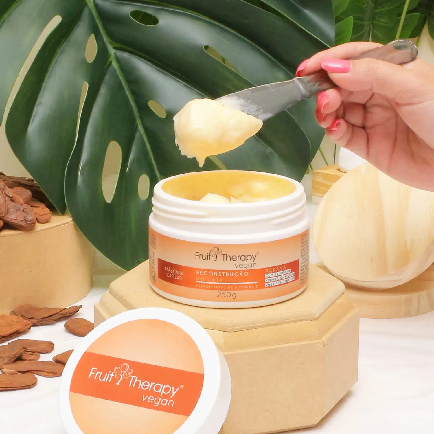 A imagem mostra a máscara de reconstrução da linha Fruit Therapy Vegan de Papaya. O pote branco com rótulo laranja está aberto. Uma mão está retirando o conteúdo amarelado e cremoso com uma espátula. Ao redor, vemos folhas verdes tropicais.