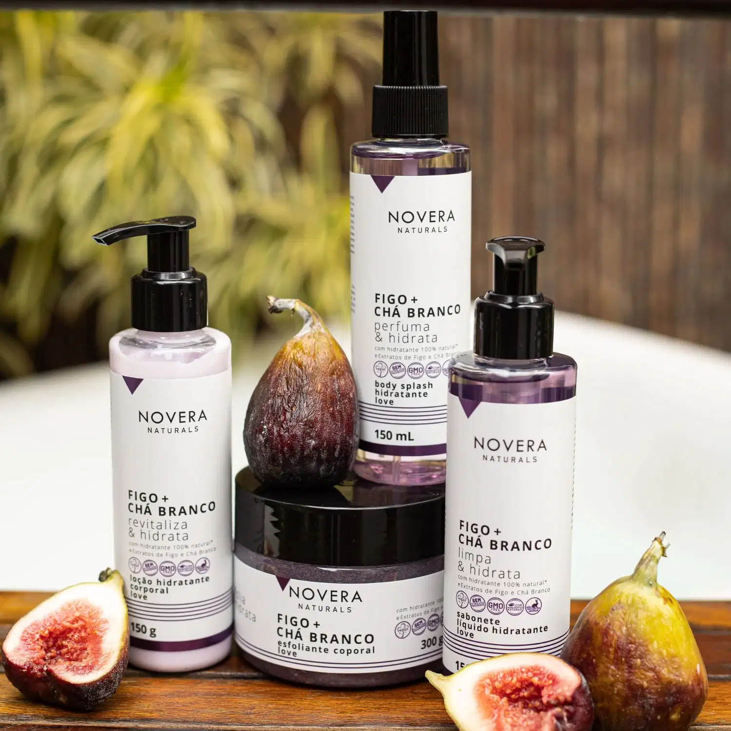 A imagem traz a linha Novera Naturals Figo + Chá Branco. São 4 itens em embalagens de plástico transparente com tons de lilás e rótulo branco. Traz esfoliante, sabonete líquido, loção corporal e body splash sobre uma superfície de madeira com figos.