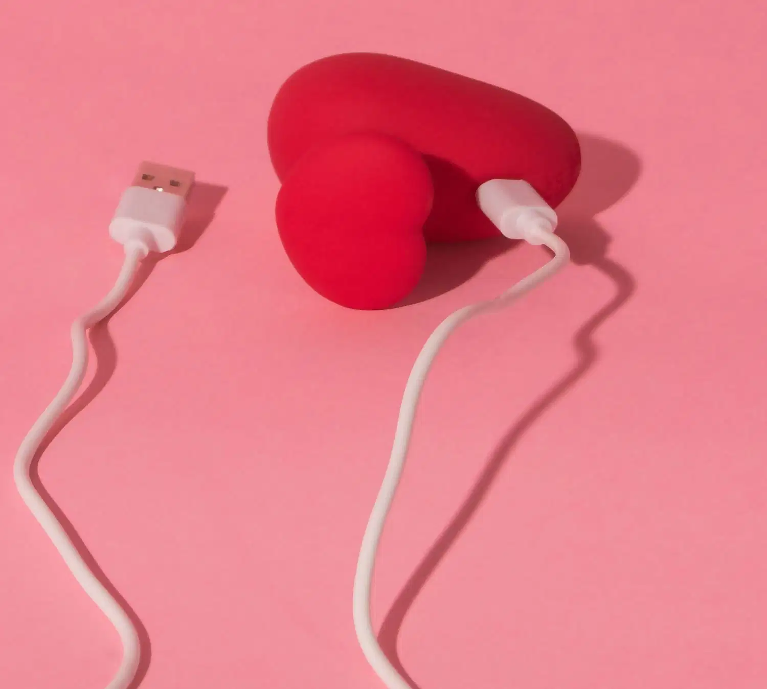 Acessório em formato de coração vermelho com cabo de carregamento USB branco, em fundo rosa sólido. Produto da marca Désir Atelier.