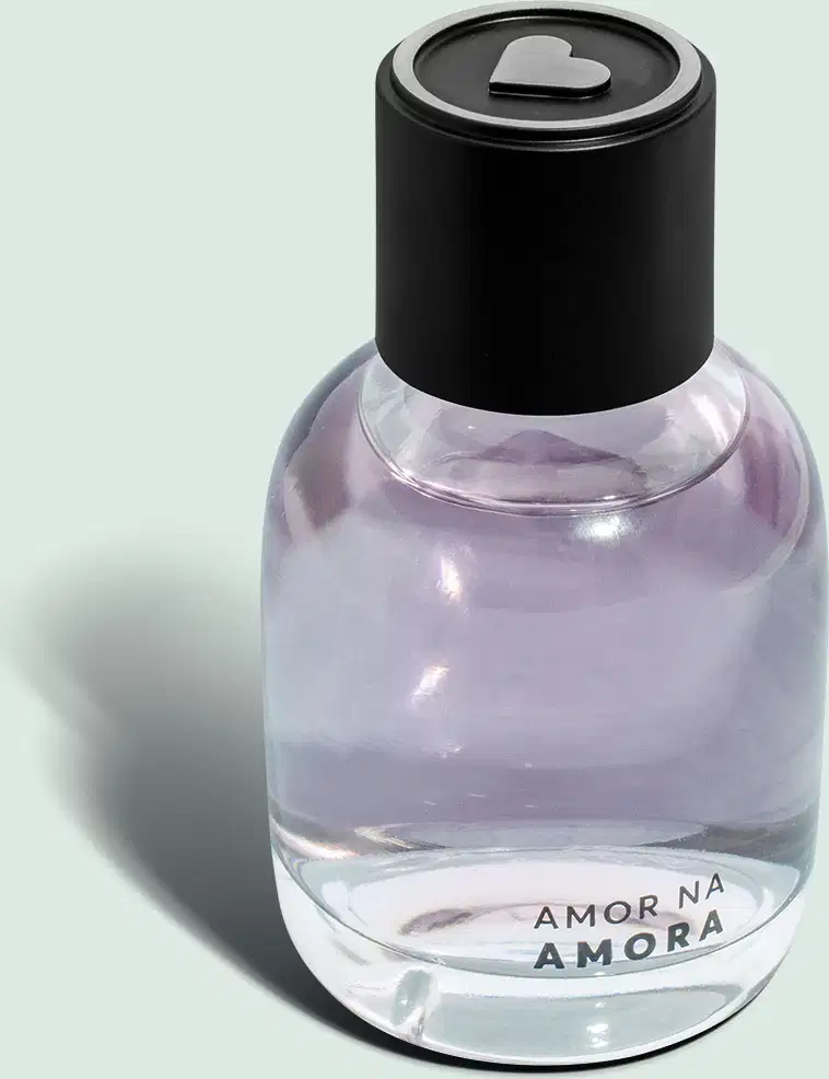 O frasco tem uma tampa preta arredondada. Na parte inferior do frasco, está impresso o texto "AMOR NA AMORA" O produto apresenta uma aparência minimalista, com o frasco transparente permitindo visualizar o líquido lilás em seu interior.