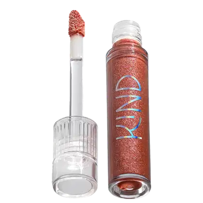 A imagem mostra a tampa da embalagem do gloss com o aplicador virado pra cima, semelhante a uma tampa de batom, transparente, cilíndrica, com uma amostra de cor. Ao lado, o frasco do gloss, com o produto de cor marrom e palavra KIND escrito na embalagem.