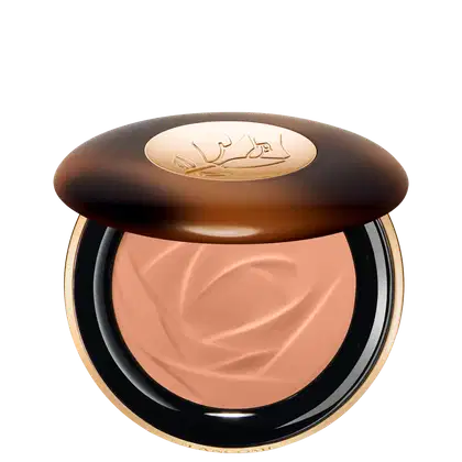 A imagem traz um sérum bronzeador compacto em uma embalagem. A tampa superior é marrom com alguns detalhes mais claros e tons mais escuros. O logotipo da marca aparece gravado em dourado no centro da tampa e o produto possui uma cor rosada.