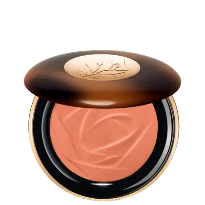 A imagem traz um sérum bronzeador compacto em uma embalagem. A tampa superior é marrom com alguns detalhes mais claros e tons mais escuros. O logotipo da marca aparece gravado em dourado no centro da tampa e o produto possui uma cor rosada.