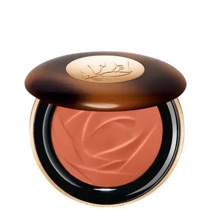A imagem traz um sérum bronzeador compacto em uma embalagem. A tampa superior é marrom com alguns detalhes mais claros e tons mais escuros. O logotipo da marca aparece gravado em dourado no centro da tampa e o produto possui uma cor rosada.