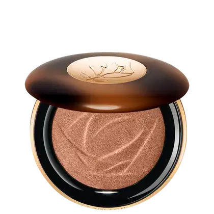 A imagem mostra o sérum iluminador Lancôme. A embalagem é redonda com tampa marrom e logotipo dourado no centro. A parte inferior preta com aro dourado. Interior contém o pó iluminador marrom com padrão rendado.