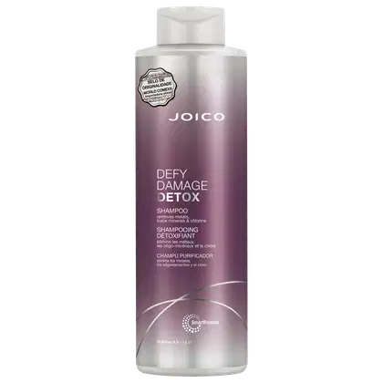 Shampoo detox Defy Damage da marca Joico. Embalagem cilíndrica roxa com detalhes prateados. Rótulo indicando que remove metais, minerais traço e cloro dos cabelos. Volume de 1L.