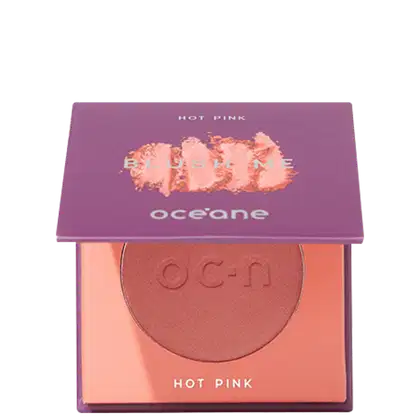 A imagem mostra o produto Blush Me da marca Océane. É uma embalagem quadrada de cor roxa que está semiaberta mostrando a cor do blush em um tom rosa com leve brilho perolado. A parte interna da embalagem é de cor pêssego claro,
