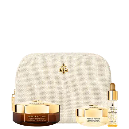 Conjunto de itens de perfumaria em embalagem bege com detalhes dourados. Inclui um frasco de creme de tratamento de mel e um frasco de óleo de tratamento, ambos com tampas douradas. Há também uma bolsa de tecido bege com um logotipo dourado.