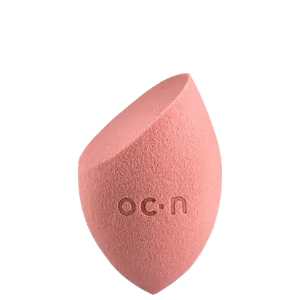 Esponja de maquiagem de cor rosa, com formato assimétrico e textura macia, com o logotipo "O.C.N." impresso na superfície.