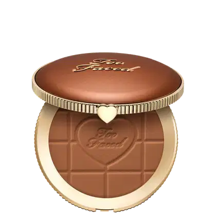 Pó bronzeador compacto com acabamento aveludado em embalagem redonda de metal dourado com detalhes em relevo e coração no centro. Apresenta textura e tonalidade marrom escura com inscrição "Too Faced" na superfície.