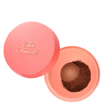 Pó solto Too Faced aberto sobre fundo branco. À esquerda, a tampa redonda e rosada exibe "Too Faced" em letras douradas. À direita, a base rosada mostra o pó solto em tom marrom-escuro ao redor do dosador "Peach Pit™"