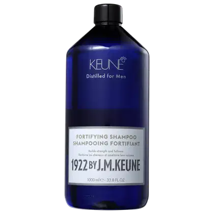 Frasco de shampoo fortificante da marca Keune, na cor azul escuro, com rótulo destacando o ano de 1922 e a frase "Distilled for Men". O produto promete construir força e volume nos cabelos.