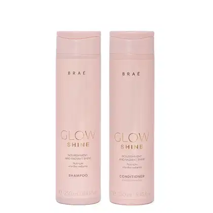Dois frascos de produtos Braé da linha Glow Shine, um de shampoo e outro de condicionador, com embalagens rosadas e textos em dourado indicando os nomes dos produtos.