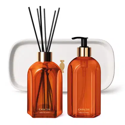 Dois vidros na cor laranja da marca Casa 214 Vanilla Sublime: um difusor de ambientes com varetas e um sabonete líquido com bico dosador preto, acompanhados de uma bandeja de lavabo branca com logo da marca O Boticário em dourado.