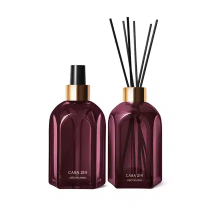 A imagem mostra dois vidros retangulares roxos, com rótulo em dourado. Um deles é o aromatizador de ambientes com válvula spray preta e o outro é um difusor com tampa dourada que acompanha varetas de madeira. Ambos são a linha Casa 214 Figo Magnífico.